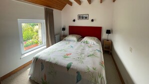 A bedroom with a wooden ceiling, a large window with a view of greenery, a bed with a floral bedspread, two bedside tables with lamps, and a red headboard. - Armor" apartment in 18th century house 5 min. from sandy beaches (Carnac)