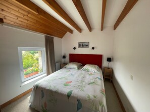 Une chambre avec un plafond en bois, une grande fenêtre donnant sur la verdure, un lit recouvert d’une courtepointe à motifs floraux, deux tables de chevet avec des lampes et une tête de lit rouge.