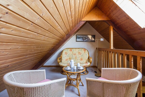Une pièce mansardée chaleureuse, avec des lambris en bois, un fauteuil à motifs floraux, des chaises en osier et une petite table sur laquelle est disposé un service à thé.