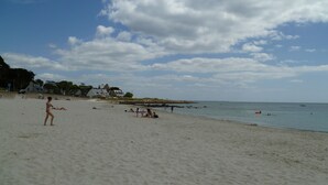 Une plage où des gens se prélassent au soleil et se baignent, quelques maisons en arrière-plan et un ciel dégagé.