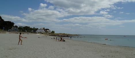 Una playa con gente tomando el sol y nadando, algunas casas al fondo y un cielo despejado.