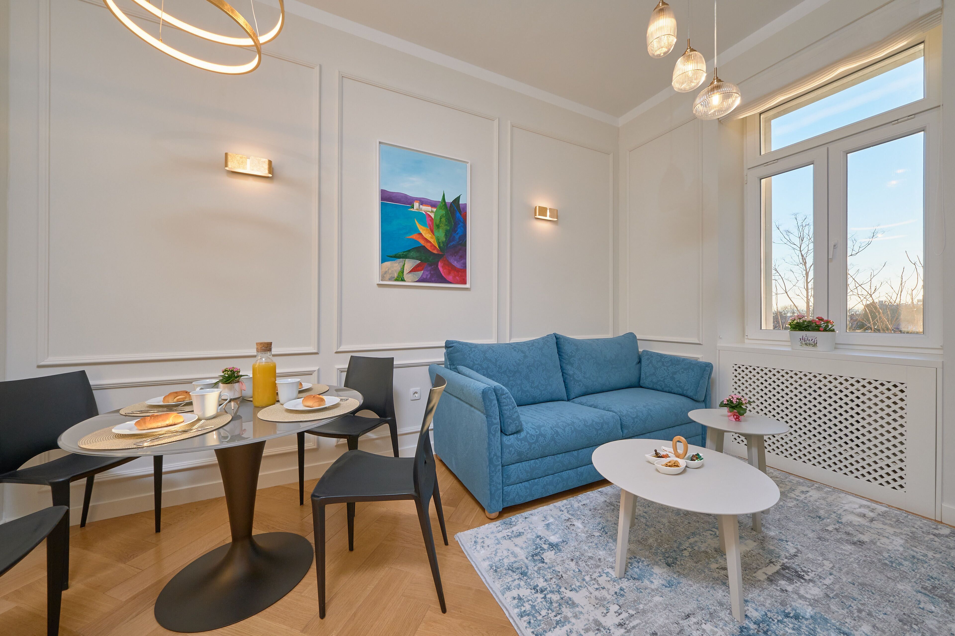 Una sala de estar moderna con un sofá azul, una mesa de comedor con sillas, una pequeña mesita auxiliar y un ventanal con vistas.