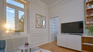 A modern living room with a large flat-screen TV, a round mirror, a small round table with decorative items, a sofa, and a window with a view of trees and the sun. - Salve Luxury Apartment by Litto (Split)