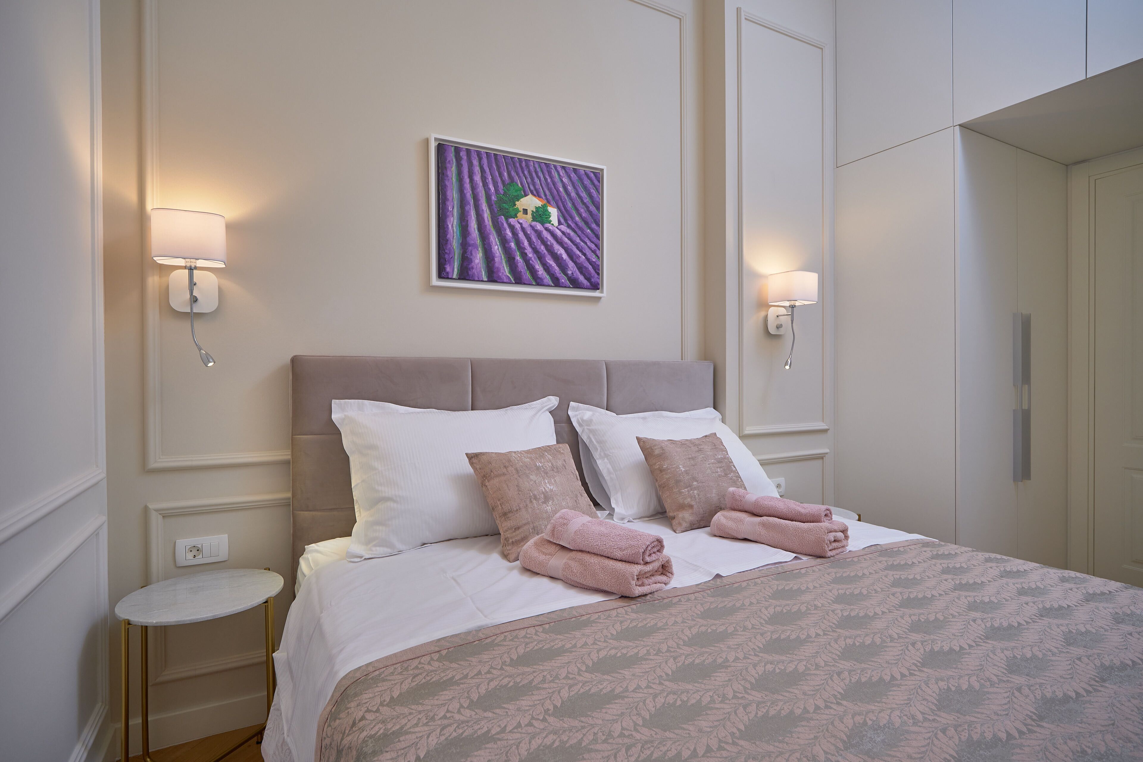 Ein ordentlich bezogenes Bett mit weißer und rosa Bettwäsche, ein kleiner runder Tisch daneben und ein an der Wand hängendes Bild von einem Haus auf einem Feld.