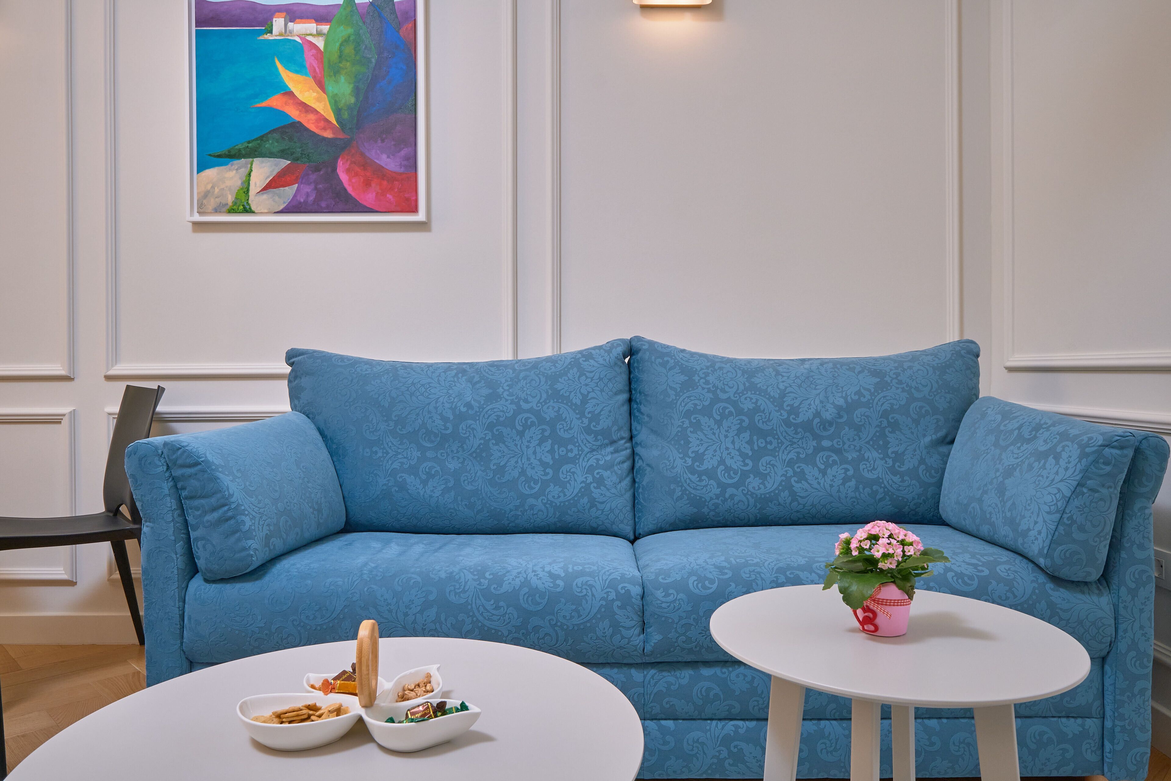 Un canapé bleu orné d’un motif, une petite table blanche sur laquelle est posé un pot de fleurs rose, et un bol blanc contenant des en-cas.