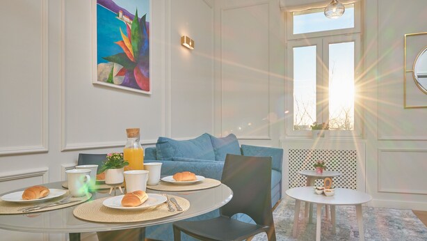 A dining area with a round table, chairs, and a sofa. There is a painting on the wall, a window with sunlight, and a small table with a plant. - Salve Luxury Apartment by Litto (Split)