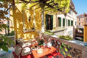 Ein Balkon mit Tisch für zwei Personen, einer Topfpflanze und Blick auf ein gelbes Gebäude mit grünen Fensterläden.
