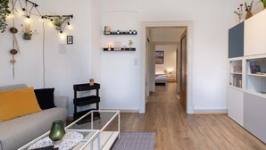 Un salon moderne avec un canapé, une table basse et des étagères. Un couloir mène à une autre pièce où se trouve un lit.