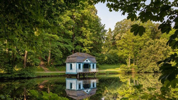 Une maison à deux étages dotée d’un balcon, entourée d’une végétation luxuriante et bordée d’une étendue d’eau paisible.