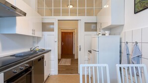 Une cuisine compacte avec des meubles blancs, une hotte en acier inoxydable et un réfrigérateur. Une porte en bois donne accès à une autre pièce.