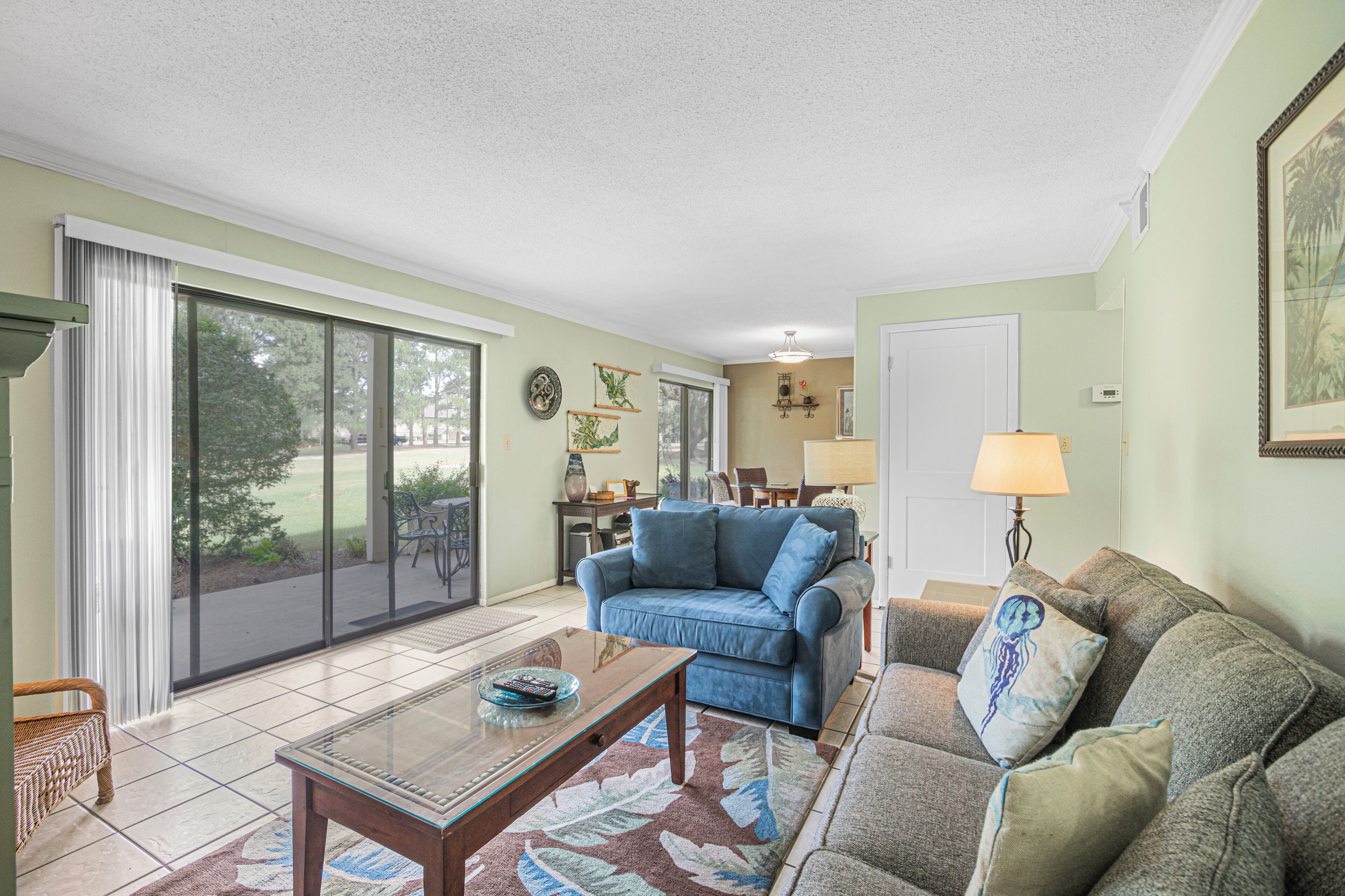 A spacious living room with a blue sofa, a grey sofa, and a glass coffee table. There is a sliding glass door leading to a patio area.