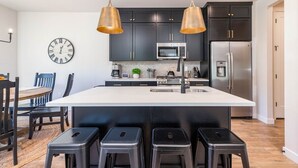 A modern kitchen with dark cabinets, stainless steel appliances, a white island with bar stools, and a wall clock.