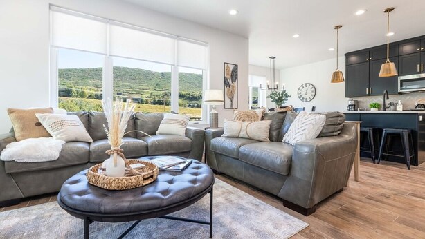 A modern living room with two grey leather sofas, a coffee table, and a view of a green landscape through large windows.