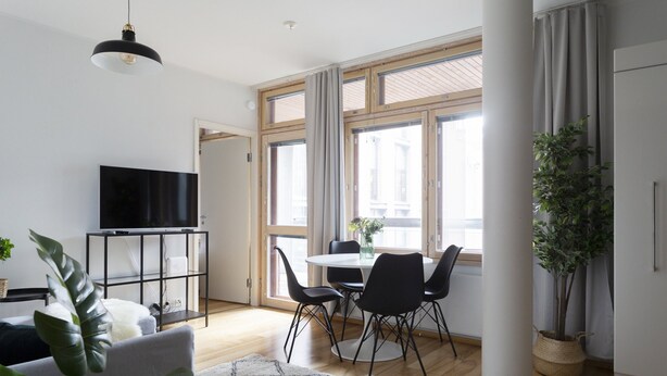 Uma sala de estar moderna com televisão de tela plana, uma área de jantar com mesa e cadeiras, e uma janela grande com cortinas.