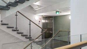 Interior moderno com escada de vidro, elevador de metal e parede branca com detector de incêndio.
