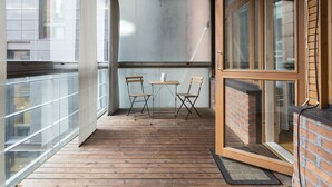 Varanda com piso de madeira, duas cadeiras, uma mesa pequena e uma porta de vidro que dá para uma parede de tijolos.