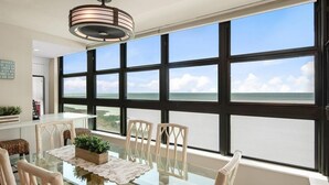 Un coin repas avec une table en verre, des chaises blanches et une vue sur l’océan grâce à de grandes fenêtres.