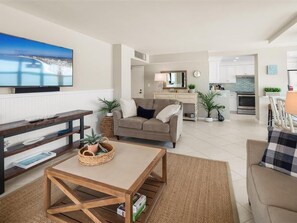 A modern living room with a sofa, coffee table, and TV. A kitchen is visible in the background.