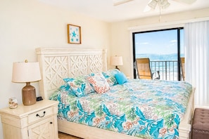 A bedroom with a bed, two bedside lamps, a nightstand, a chair by the window, and a view of the ocean.