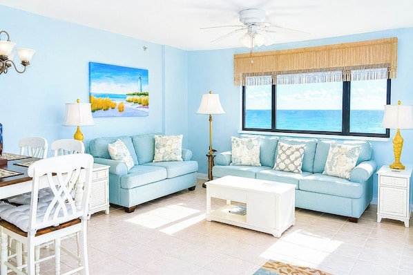 A living room with a blue sofa set, a dining table with chairs, a coffee table, and a large window with a view of the sea.