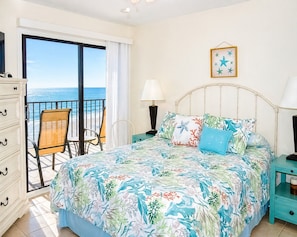 A bedroom with a bed, a chair, a nightstand, a dresser, and a view of the ocean.