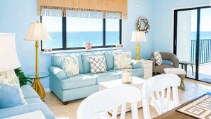 Una sala de estar con un sofá azul claro, sillas blancas, una mesa de centro, una lámpara y vista al océano a través de ventanas grandes.