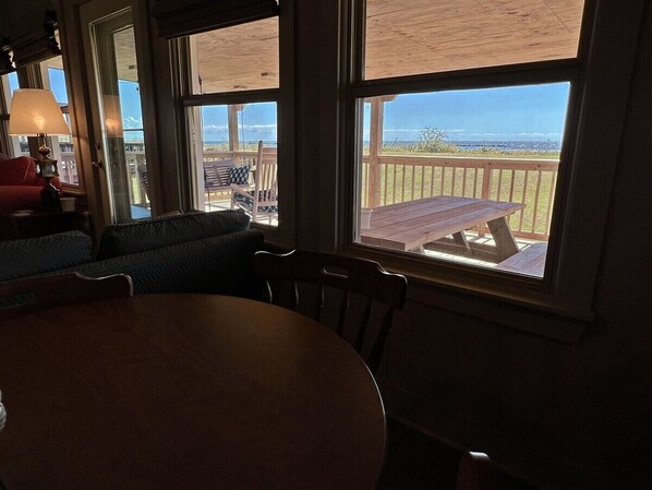 A dining area with a table and chairs, large windows with a view of a deck and the ocean, and a lamp on a side table.