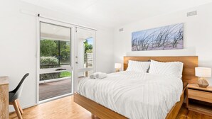 A bedroom with a large bed, a wooden headboard, a nightstand, a chair, and a sliding glass door leading to an outdoor area. - Cape Surf House in Cape Woolamai (Cape Woolamai)