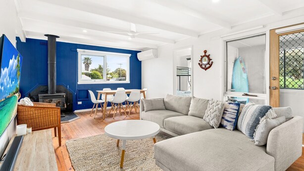 A living room with a sofa, a coffee table, and a dining area with chairs. There is a fireplace, a large window, and a door with a view of the outdoors. - Cape Surf House in Cape Woolamai (Cape Woolamai)