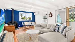 A living room with a sofa, a coffee table, and a dining area with chairs. There is a fireplace, a large window, and a door with a view of the outdoors. - Cape Surf House in Cape Woolamai (Cape Woolamai)