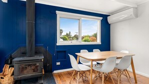 A room with a blue wall, a white window, a wooden table with white chairs, and a wood-burning stove. - Cape Surf House in Cape Woolamai (Cape Woolamai)