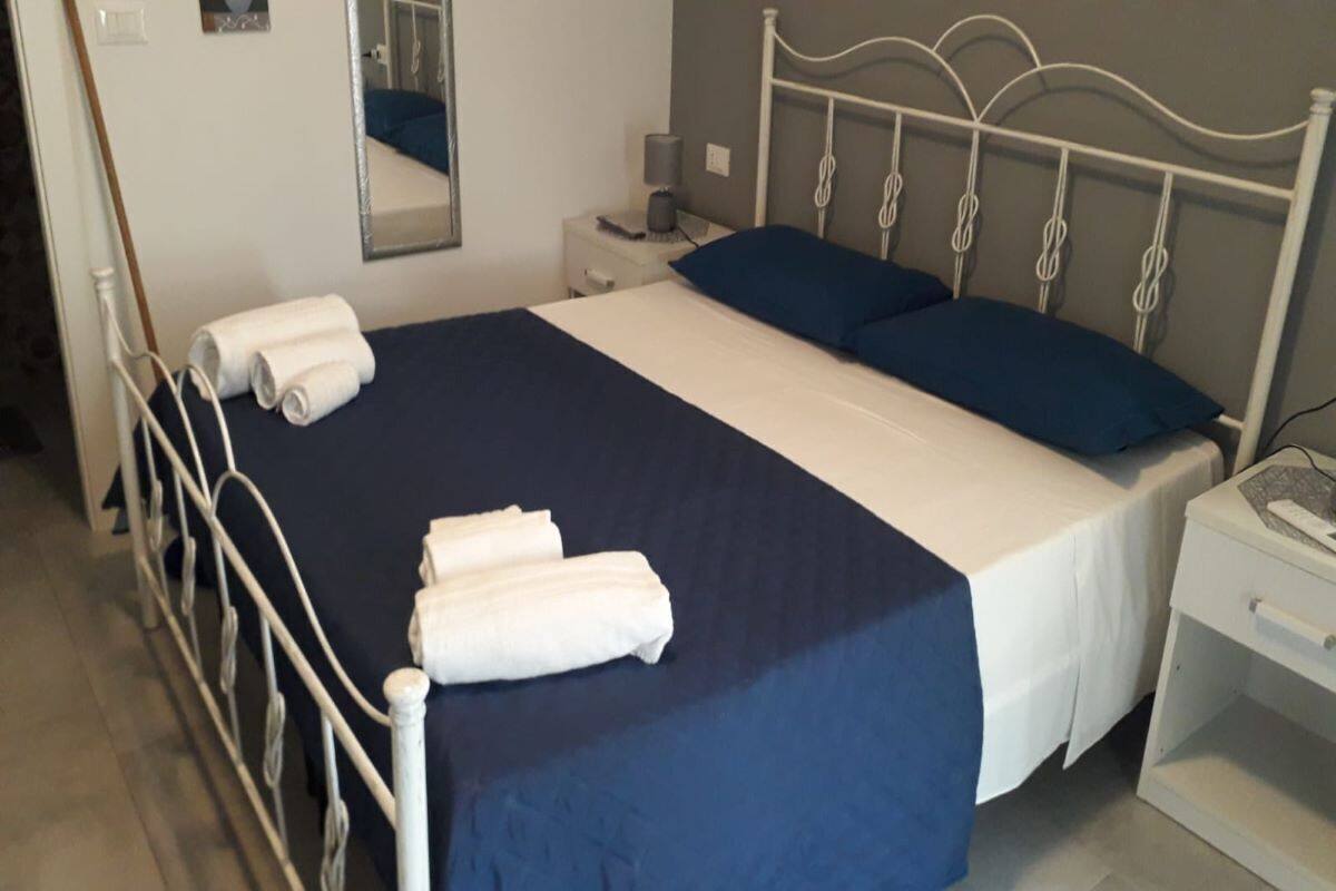 Een net opgemaakt bed met wit en blauw beddengoed, een nachtkastje met een lamp en een spiegel aan de muur.