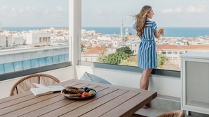Un balcon donnant sur une ville côtière, une table avec des fruits et un livre, et une personne tenant un verre.