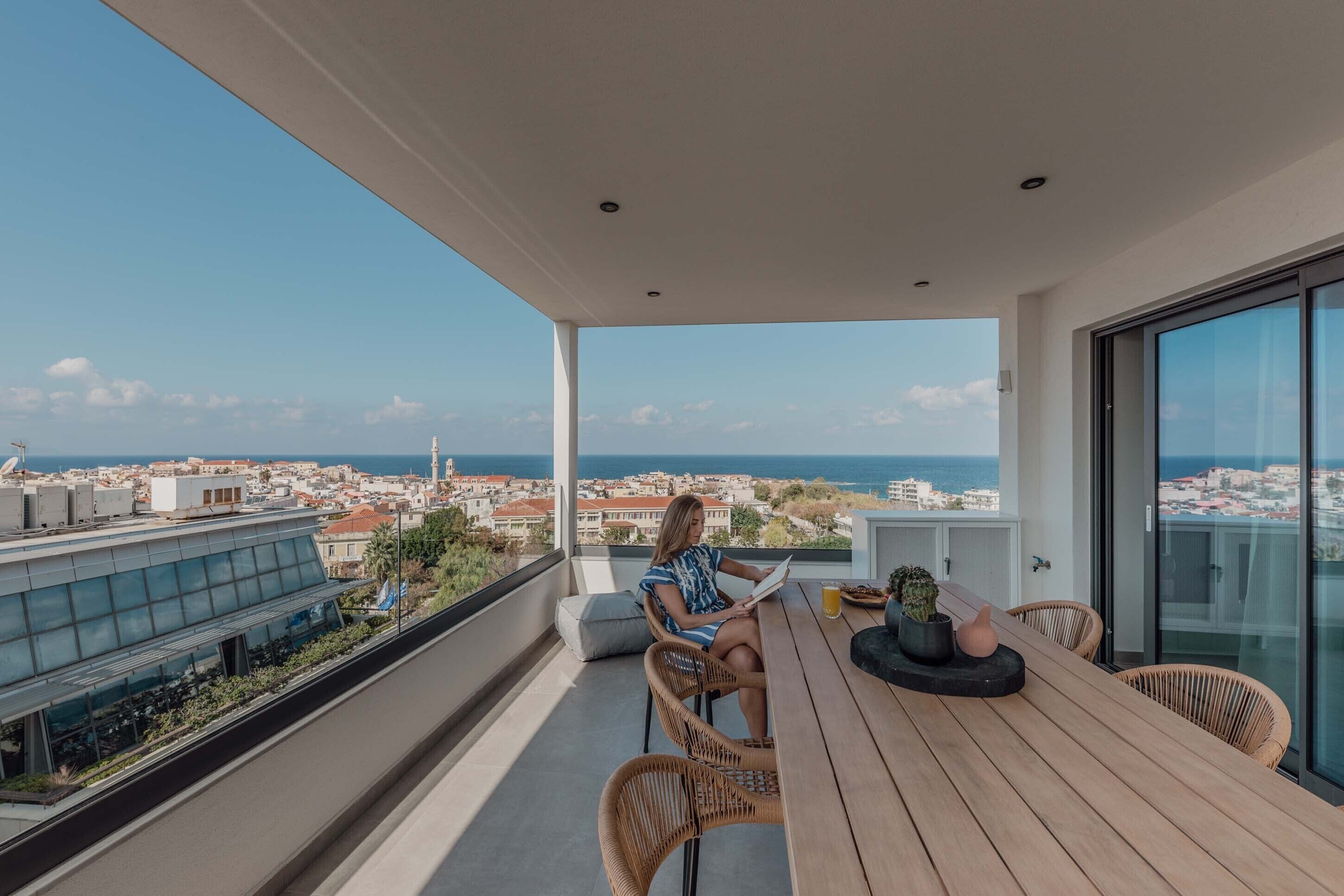 A balcony with a view of the city and sea, a wooden table, wicker chairs, and a person reading a book.