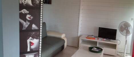 A living room with a grey sofa, a white coffee table, a TV on a white stand, and a floor fan.