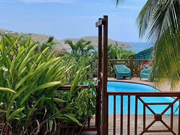 A wooden deck leading to a pool with a view of the ocean and hills in the background.