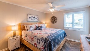Ein Schlafzimmer mit Bett, Deckenventilator, Fenster mit Jalousien, Nachttisch mit Lampe und Wanduhr.