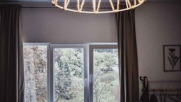 Ein Zimmer mit großem Fenster, Vorhängen, einer Hängelampe und einem gerahmten Bild an der Wand.