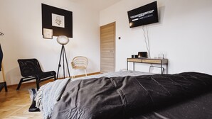 Ein Schlafzimmer mit einem Bett, einem Schreibtisch mit Laptop, einem Sessel, einer Stehlampe und einem Fernseher an der Wand.