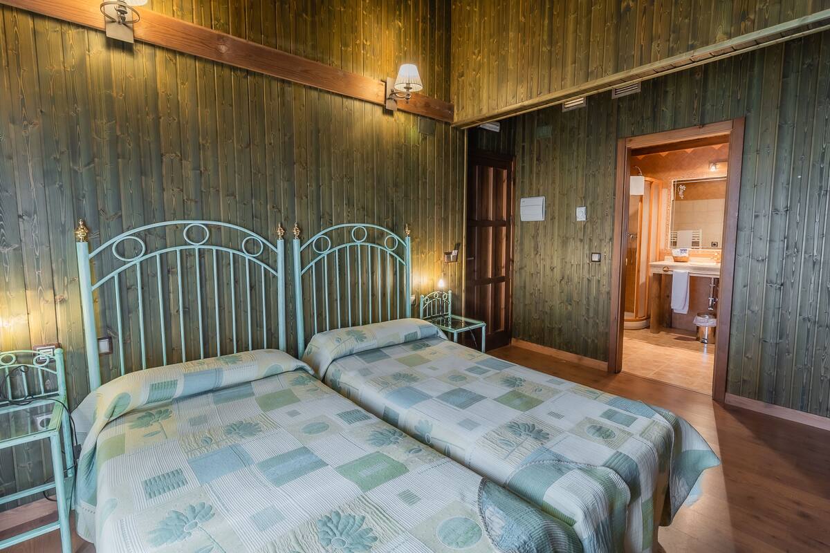 A room with two beds, wooden walls, a wooden ceiling, and a bathroom visible through an open door.