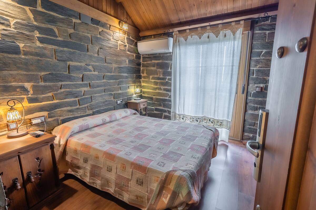 A bedroom with a stone wall, a bed with a patterned bedspread, a bedside table with a lamp, and a window with curtains.