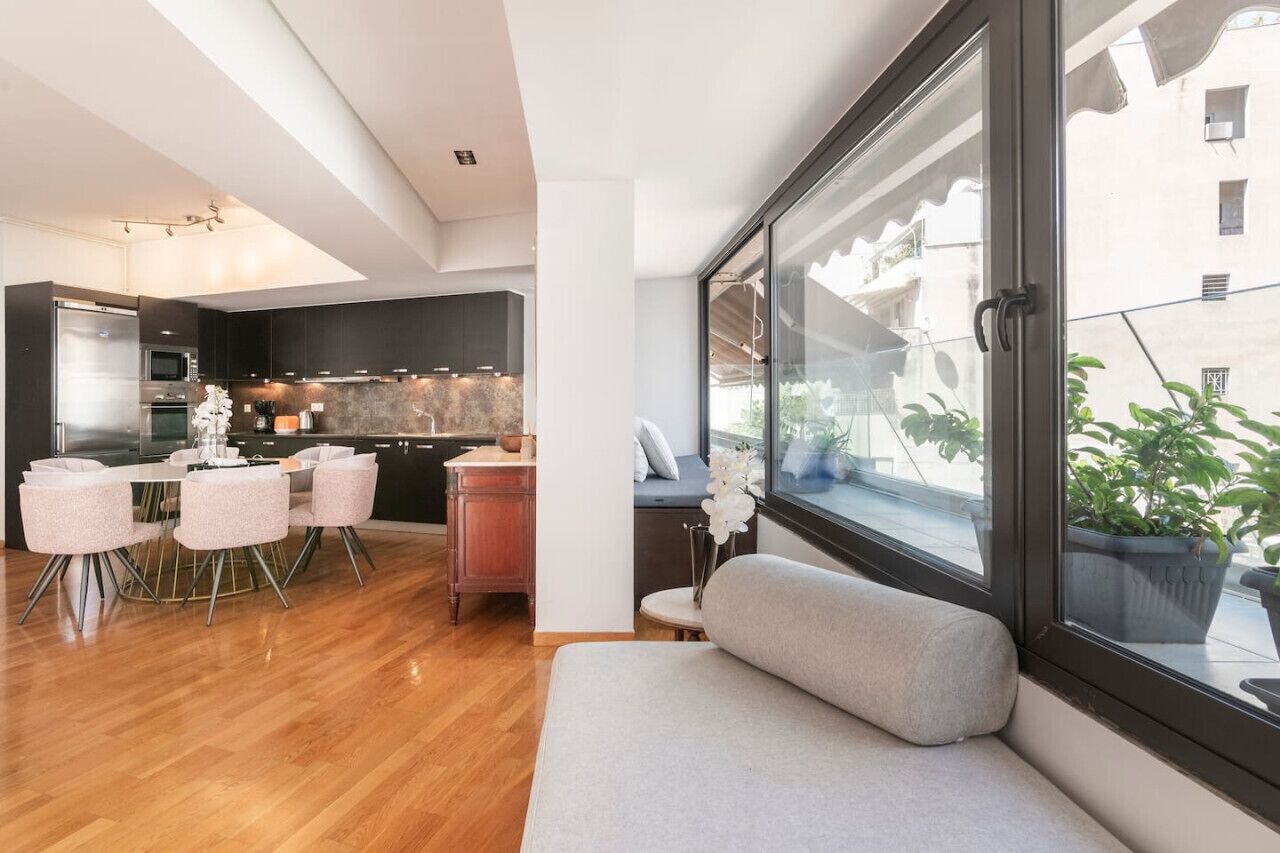 Een moderne keuken met donkere kasten, een zithoek met een ronde tafel en stoelen, en een groot raam met uitzicht op gebouwen.