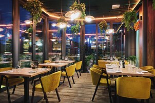 A restaurant interior with wooden tables, yellow chairs, hanging plants, and large windows.