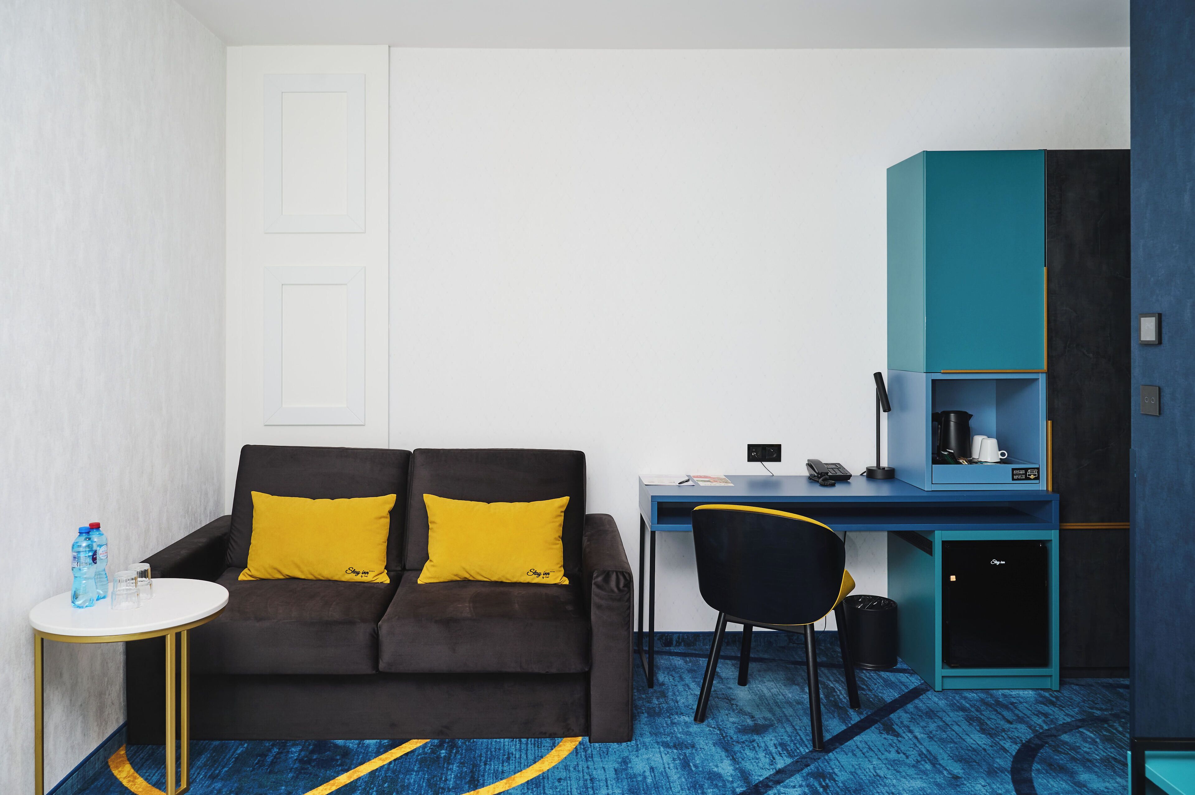 A compact room with a dark brown sofa, a small round table, a desk with a chair, and a kitchenette area.