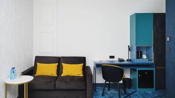 A compact room with a dark brown sofa, a small round table, a desk with a chair, and a kitchenette area.
