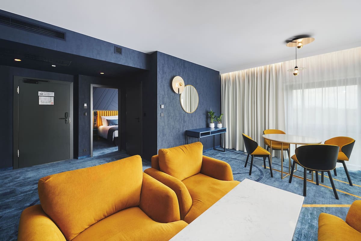 Ett modernt hotellrum med en blå accentvägg, en rund spegel, en matplats med bord och stolar samt en soffa.