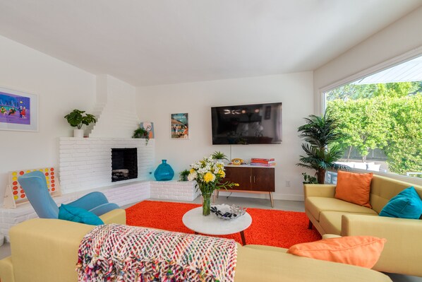 Een moderne woonkamer met een wit bakstenen haardvuur, een flatscreen televisie en een groot raam met uitzicht op bomen.