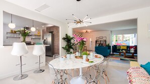 A modern kitchen with white cabinets, a marble island, and pendant lighting. There is a dining area with a round table and chairs.