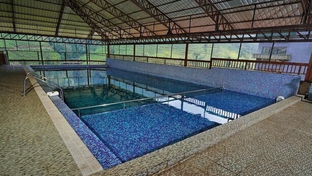 Una piscina interior con agua azul cristalina, rodeada por un muro de azulejos y una estructura de techo metálico.