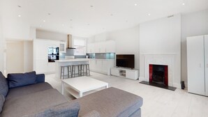 Ein modernes Wohnzimmer mit grauem Sofa, einem weißen Couchtisch, einem Fernseher, einem Kamin und einer Küchenzeile mit weißen Schränken und einem Barhocker.